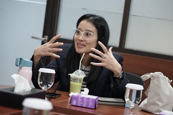 kolaborasimarcellazalianty4 dompet dhuafa Woman in a dark blazer and white-framed glasses speaks animatedly at a meeting, hands spread in gesture, seated at a table with drinks and tissues nearby.