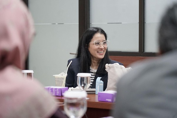 kolaborasimarcellazalianty5 dompet dhuafa Smiling woman with glasses sits at a conference table during a meeting, with drinks and purple tissue boxes nearby.