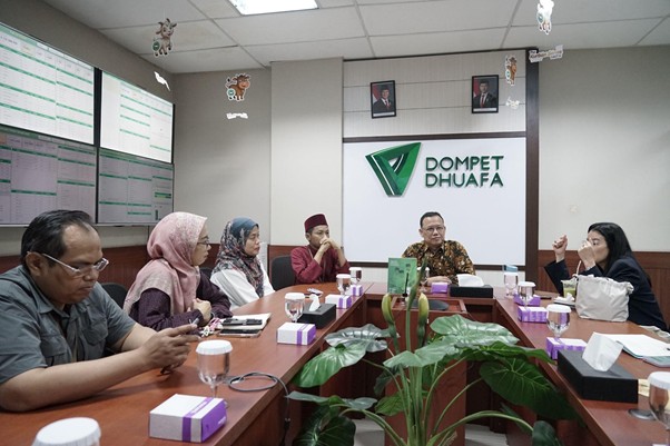 kolaborasimarcellazalianty7 dompet dhuafa A group of eight people sits around a conference table in a meeting room, several with notebooks and cups, with a Dompet Dhuafa sign on the wall behind them and potted plants in the foreground.