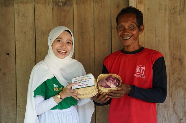 kurban rois ojk 1 dompet dhuafa Smiling woman in a white hijab and green shirt hands a wicker basket of bread to a man in a red shirt, against a wooden wall.