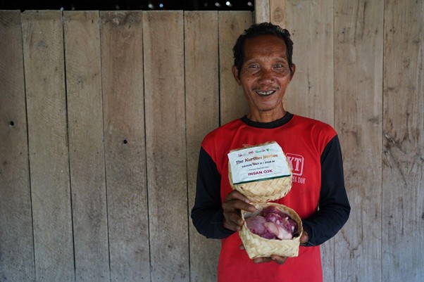 kurban rois ojk 10 dompet dhuafa Smiling man in a red and black shirt stands in front of a wooden fence, holding two woven baskets with produce visible in them.