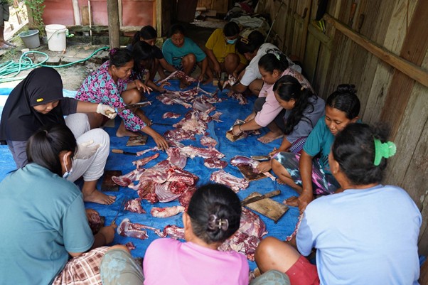 kurban rois ojk 6 dompet dhuafa Group of people sitting on a blue tarp, processing and cutting raw meat from several animal carcasses inside a simple shelter.