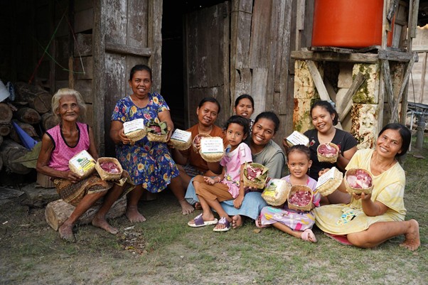 Group of women and children sitting on the yard outside a weathered wooden building, each holding a woven basket of food and smiling.