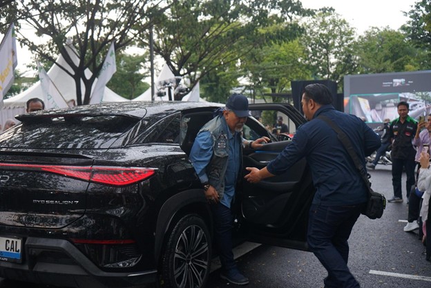 kurbanaval20263 dompet dhuafa Two men assist a man stepping out of a black SUV at an outdoor car show, with tents and onlookers in a park setting.