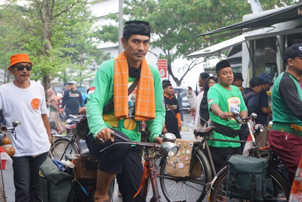 kurbanaval20264 dompet dhuafa Man in a green shirt with an orange scarf sits on a bicycle at a bike-parade or gathering, with other cyclists and onlookers in the background.