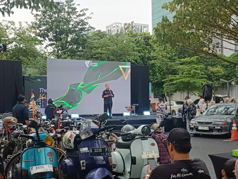 kurbanavalthekurbanseries1447h1768x576 dompet dhuafa Outdoor stage event with a man speaking on a platform in front of a large screen, motorcycles parked along the foreground, and people gathered nearby.