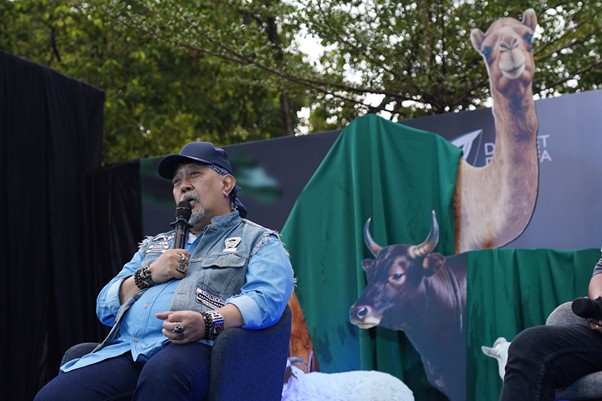 kurbanvala 2026 indro warkop 1 dompet dhuafa Man in a cap and denim shirt speaks into a microphone while seated on a stage with animal cutouts in the background.