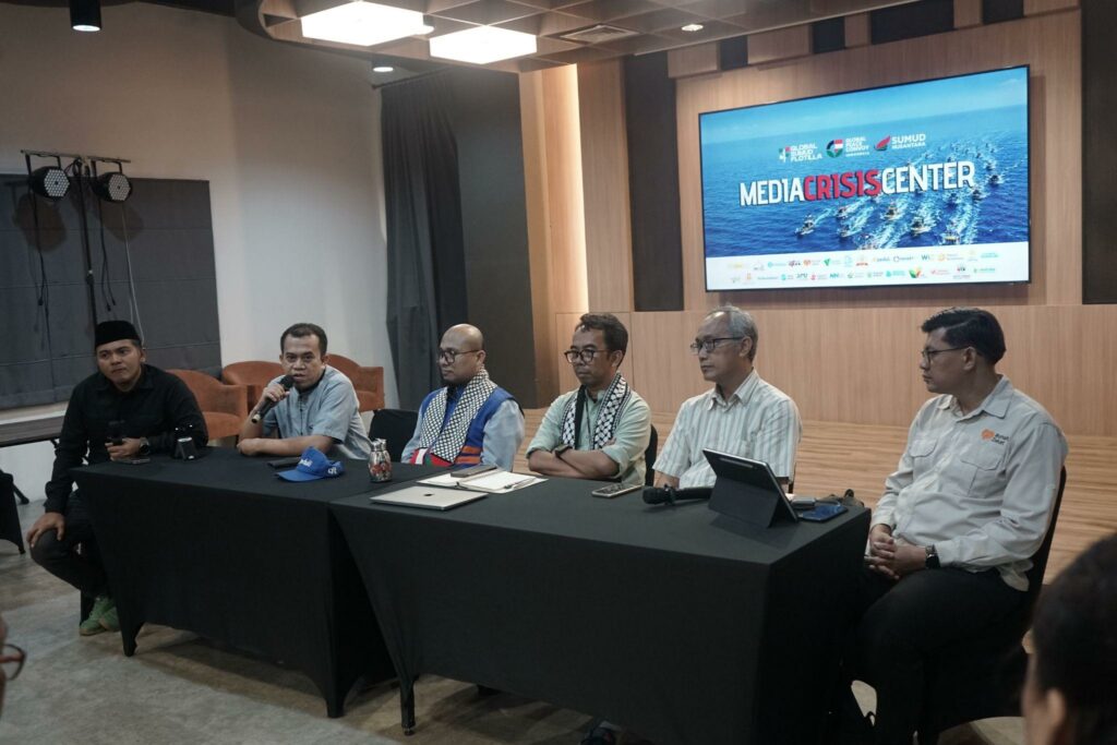 Six people seated at a long conference table with microphones; a large screen reads MEDIA CRISIS CENTER in the background.