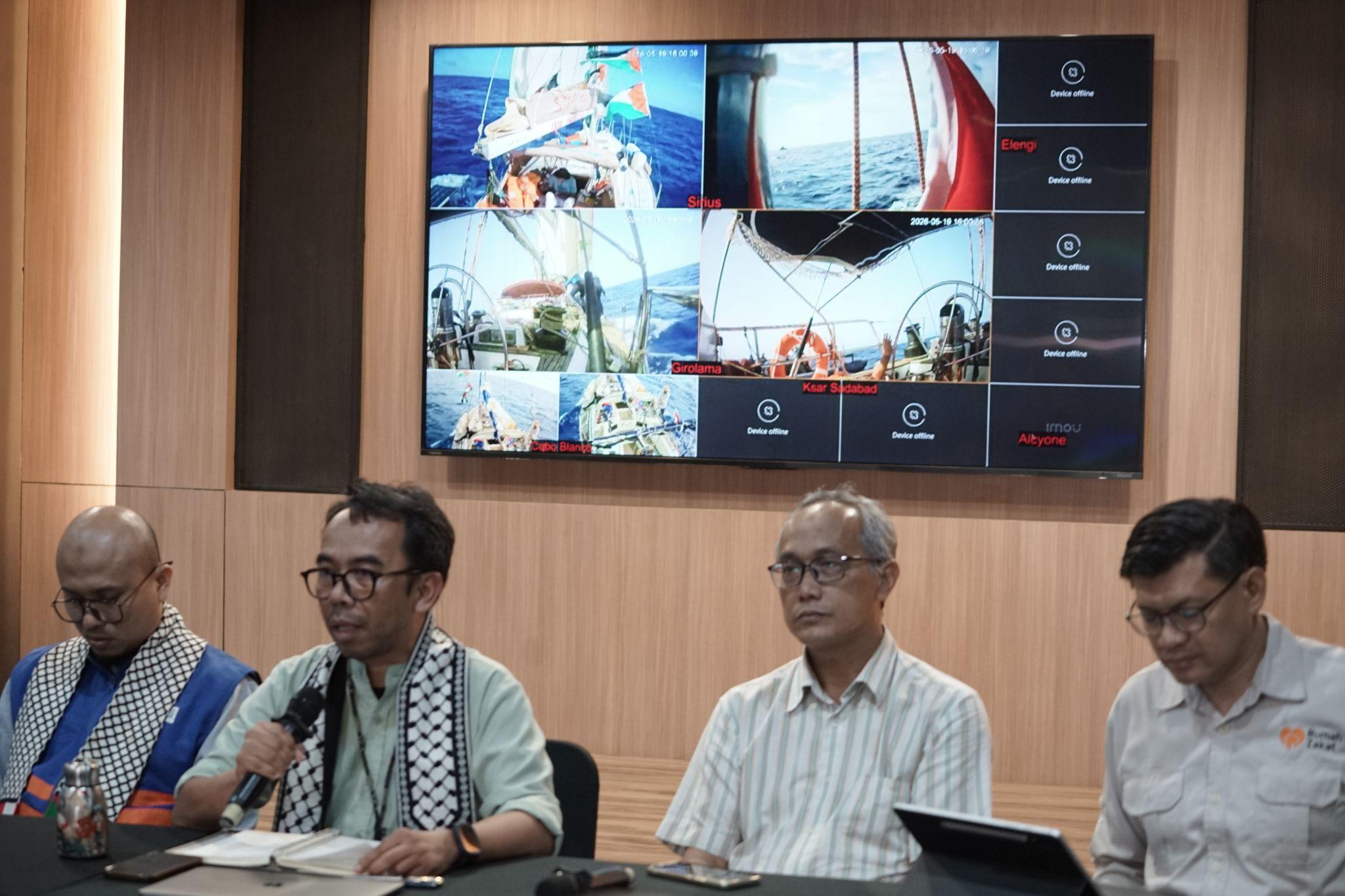 Three men sit at a conference table with microphones in front of a large video wall displaying sailing photos of yachts at sea.