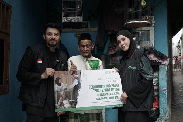 ngabuburithamidahdanfarhad12 dompet dhuafa Three volunteers in black outfits present a donation box to an elderly man in a doorway, promoting the distribution of 100 zakat fitrah packages in Bogor, West Java.