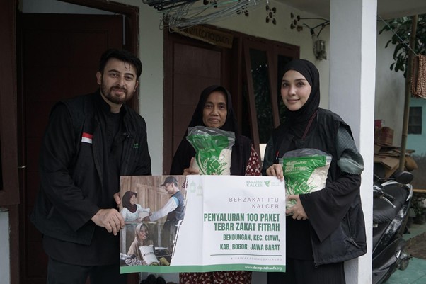 ngabuburithamidahdanfarhad4 dompet dhuafa Three people, two women wearing hijabs and a man, stand on a porch holding a banner about zakat distribution in West Java.