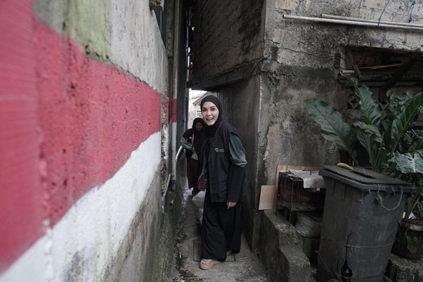 ngabuburithamidahdanfarhad7 dompet dhuafa Woman in a black hijab and abaya stands in a narrow alley between rough concrete walls, smiling at the camera with a child visible in the background.