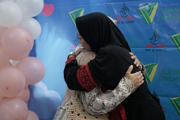 Two women wearing hijabs hug warmly at an event, standing in front of a blue banner with logos and pink balloons nearby.