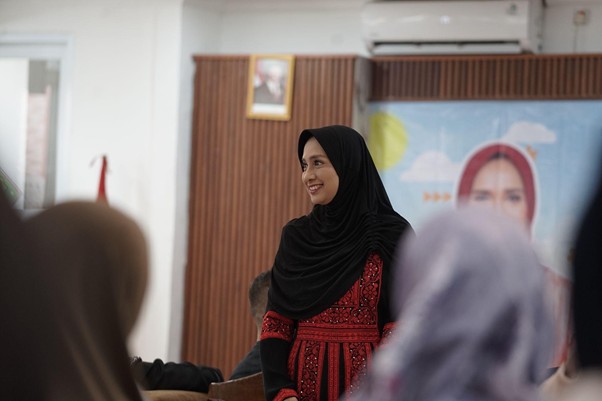 Woman wearing a black hijab and red patterned dress smiles while speaking at the front of a room with attendees in the foreground.
