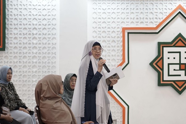 Woman in a light gray hijab speaks into a handheld microphone while holding a notebook, addressing seated women in a bright hall with decorative wall art.