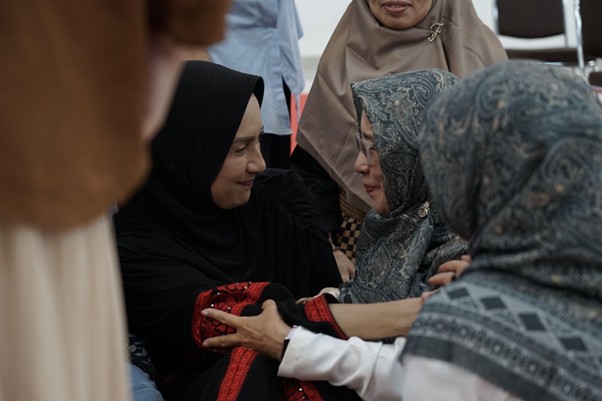 Several women in hijabs share a supportive hug, hands joined, in a close group setting.