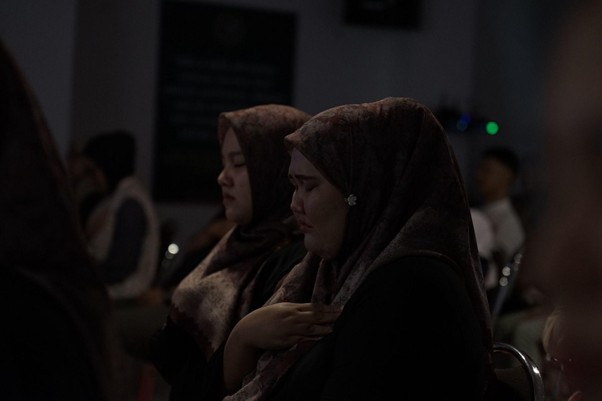 Two women wearing hijabs sit in a dim room, one with a hand over her chest during a ceremony or gathering, with others in the background.