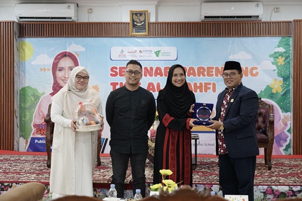 Four people stand on a stage during an awards ceremony in front of a colorful banner; from left to right: a woman in a beige hijab holding a gift basket, a man in black, a woman in a black hijab holding a glass trophy, and a man in a suit presenting the trophy.