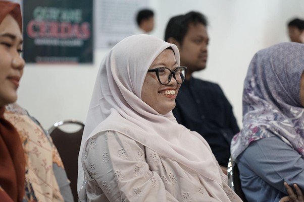 Smiling woman in a light pink hijab and glasses sits among others at an indoor event or conference.