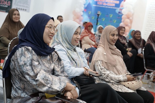 A group of women wearing hijabs seated in a hall at an event, smiling and watching a presentation, with balloons and banners in the background.