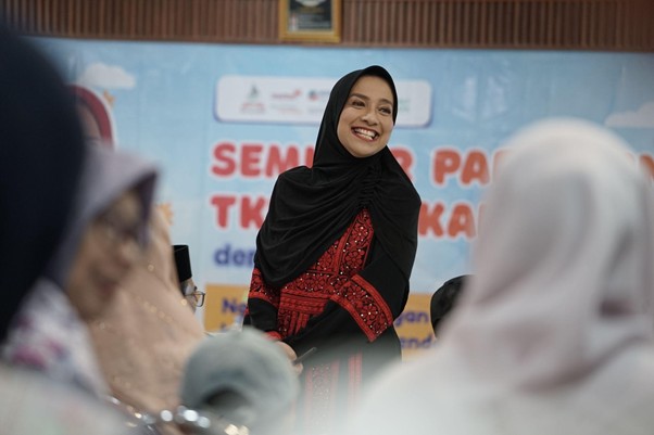 Smiling woman in a black hijab and red dress stands at an event, with a blue banner in the background and blurred audience members in the foreground.