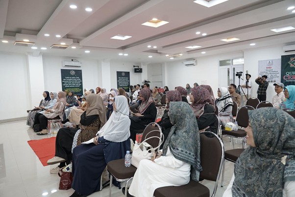 Group of women wearing hijabs seated in a conference hall listening to a presentation on stage unseen from this angle (audience shot).