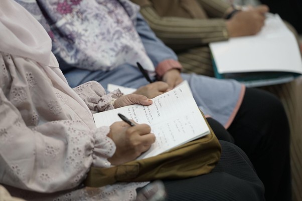 Person in a pink patterned sleeve writing in a notebook at a group meeting or class.