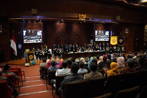 pelantikanagustinozulys4 dompet dhuafa Stage of a graduation ceremony: graduates in gowns and caps seated at a long table on stage, audience in seats facing them, Indonesian flag on the left, and screens showing the proceedings.