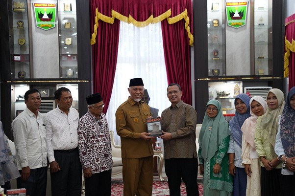 pemprovsumaterabarat10 dompet dhuafa Group of people in formal attire stand in a ceremonial room with red drapes as two men at center exchange a book or award; others look on and smile.