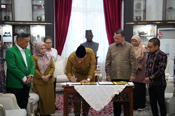 pemprovsumaterabarat5 dompet dhuafa Officials observe as a man signs a document on a wooden table during a formal ceremony in a decorated room.