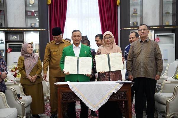 pemprovsumaterabarat7 dompet dhuafa Two officials hold signed documents at a formal ceremony, surrounded by colleagues in a decorated room with red curtains and a lace-covered table.