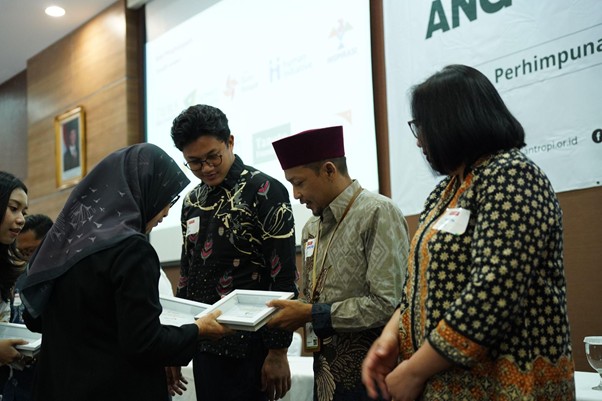 penghargaanpenghimpunanfilantropiindonesia2 dompet dhuafa A woman in a hijab receives a boxed award from a man in batik shirt on a stage, with two others watching in a conference room.