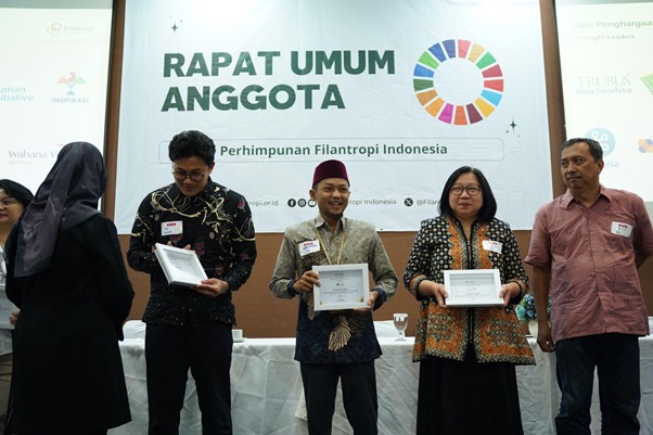 penghargaanpenghimpunanfilantropiindonesia3 dompet dhuafa Five people stand on a stage during an awards ceremony, each holding a framed certificate; a banner behind reads 'RAPAT UMUM ANGGOTA' with a colorful circular logo.