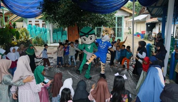 Children form a circle around two costumed mascots (green ninja and tiger) performing under a blue canopy, with families watching.