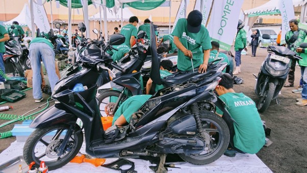 Group of technicians in green shirts repairing motorcycles under a canopy at an outdoor event, with several bikes on stands and people working on them.