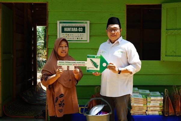 Man in a white shirt and cap hands a green certificate to a woman in a brown hijab outside a green building, with boxes nearby.