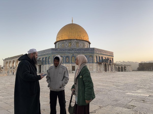 Three people stand on the stone courtyard in front of the Dome of the Rock, talking at sunset/sunrise in Jerusalem.