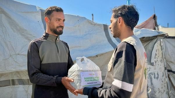 Two men exchange a bag of aid in front of tented shelters, both smiling, in an outdoor camp setting.
