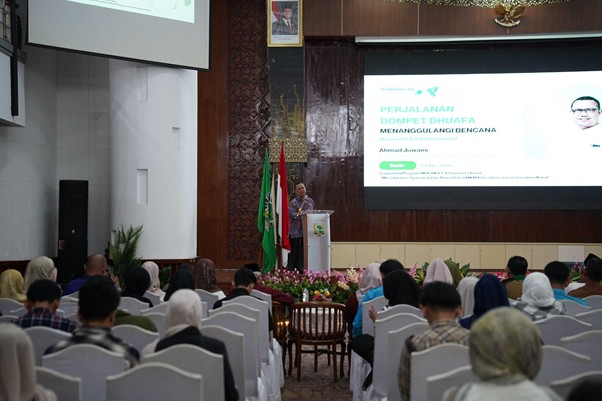 programmufakat5 dompet dhuafa Speaker at a podium presenting to an audience in an auditorium; a large projection screen shows a slide about Dompet Dhuafa and disaster relief, with flags and flowers nearby.