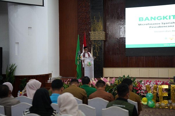 programmufakat7 dompet dhuafa Speaker at a podium presenting to an audience in a conference hall, with flags behind him and a large presentation screen to the right; the stage is decorated with flowers and balloons.