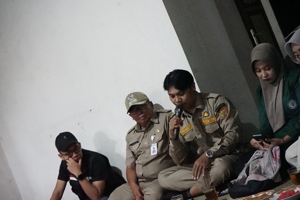 Group of people seated on the floor in a small briefing; two men in khaki uniforms, a man in a black cap, and a woman in a hijab nearby.