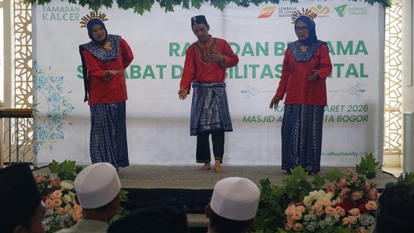 Three performers in red tops and blue traditional outfits on stage, performing at a Ramadan event with a banner behind and floral decor in front.