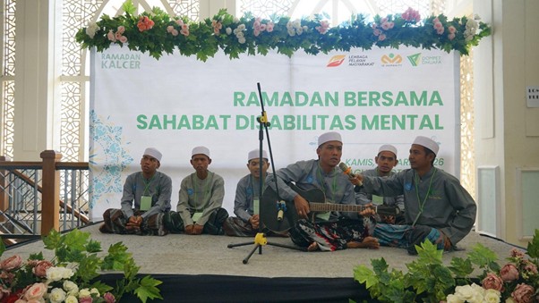 Group of men in traditional attire wearing white caps perform on a stage during a Ramadan event for people with mental disabilities, with a banner reading 'RAMADAN BERSAMA SAHABAT DIABILITAS MENTAL' and floral decorations in front.