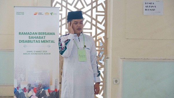 Man in white traditional clothing and a black cap speaks into a microphone at a Ramadan event for friends with mental disabilities, standing beside a banner that reads 'Ramadan Bersama Sahabat Disabilitas Mental'.