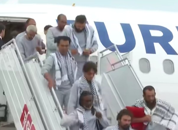 Group of men disembarking from a white airplane via mobile stairs, with blue lettering visible on the fuselage in the background.