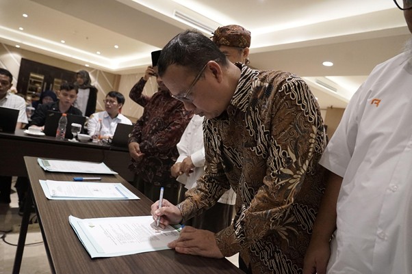sklaz20266 dompet dhuafa Man in a batik shirt signs a document at a table as colleagues observe in a conference room.
