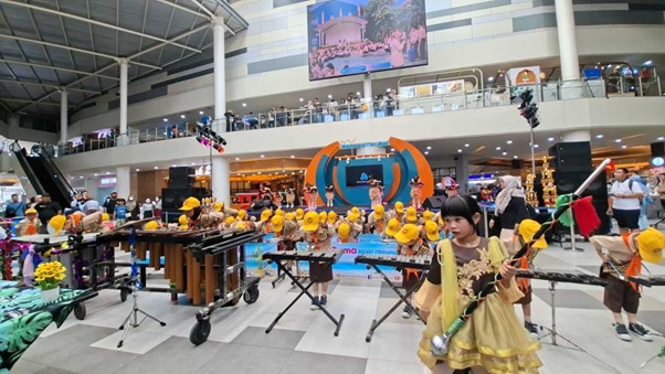Children wearing yellow hats perform music with keyboards and percussion on a mall stage, a girl in a beige dress plays a string instrument in the foreground.