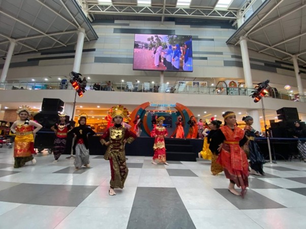 Group of dancers in vibrant traditional costumes performing a cultural dance on a mall stage, with a backdrop arch and audience on a balcony above.