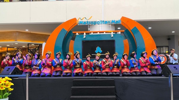 Dancers in bright traditional costumes perform a synchronized routine on an outdoor stage, with orange and teal arch structures and a 'Metropolitan' backdrop sign behind them.