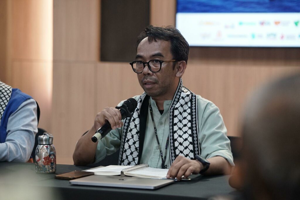 Man speaking into a handheld microphone at a panel, with a notebook and water bottle on the table in front of him.